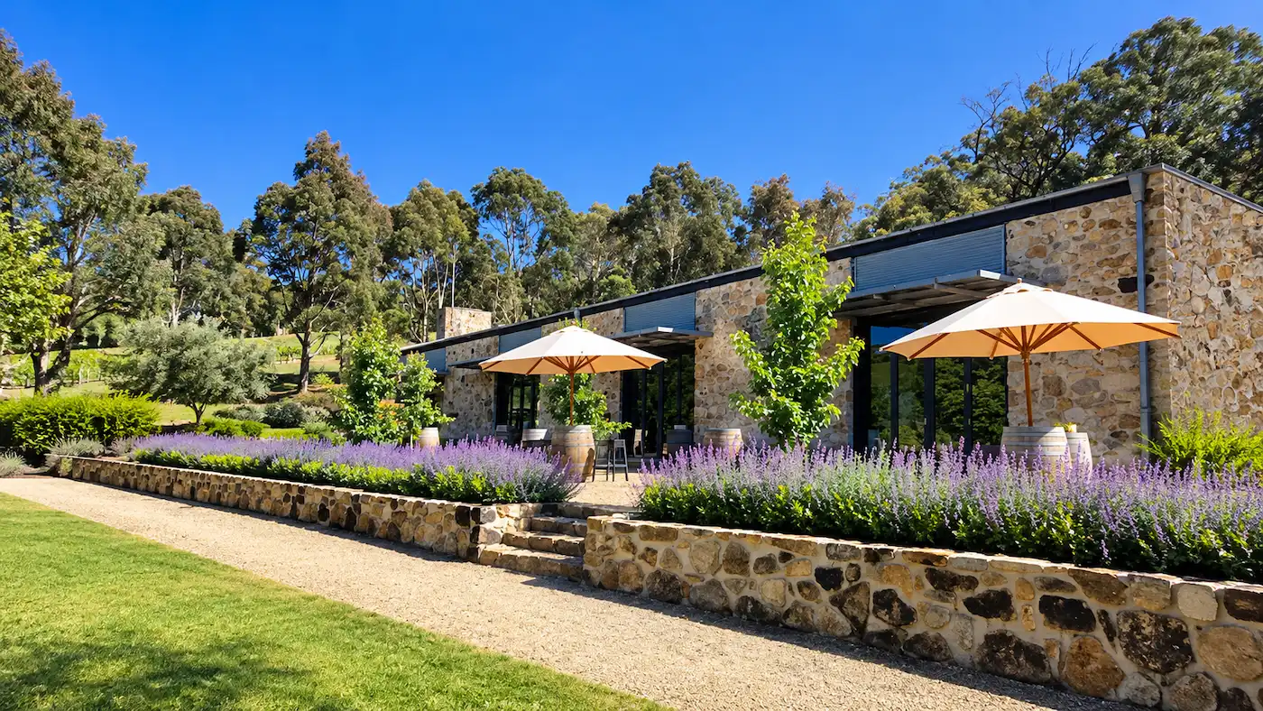 Sunlit stone winery with a landscaped garden terrace, outdoor seating beneath cream umbrellas, purple flowering plants, and surrounding trees